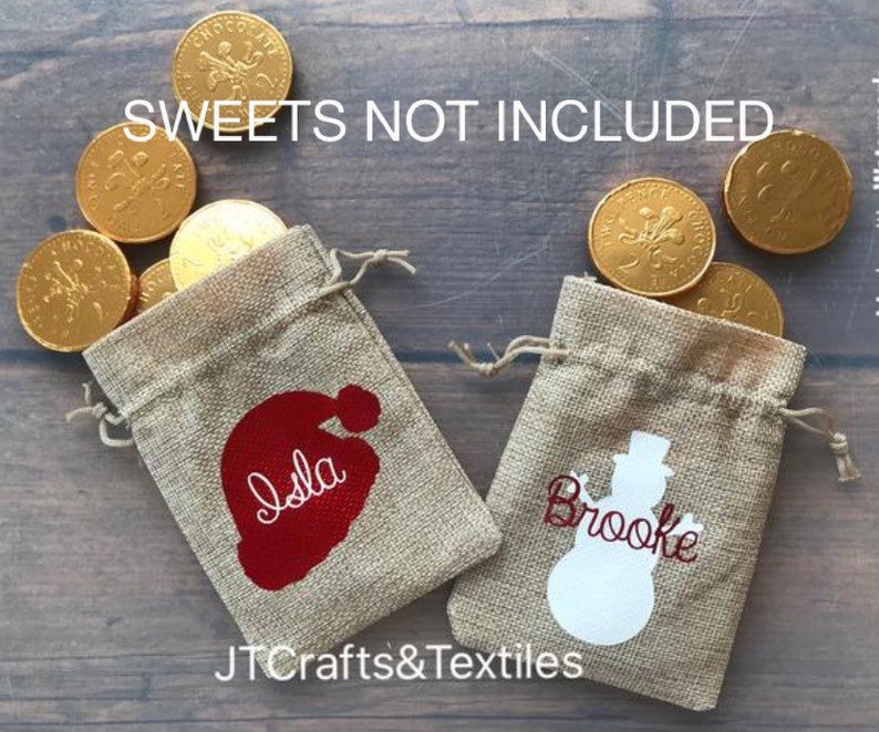 May include: Two small burlap gift bags with personalized Christmas designs. The bag on the left has a red Santa hat with the name "Isla" written in white. The bag on the right has a white snowman with the name "Brooke" written in red. The text "SWEETS NOT INCLUDED" is visible at the top of the image.