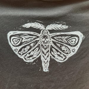 May include: A black t-shirt with a white print of a stylized moth with a skull in the center.