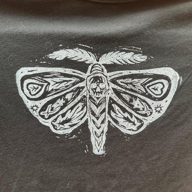 May include: A black t-shirt with a white print of a stylized moth with a skull in the center.