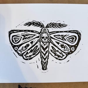May include: Black and white linocut print of a moth with a skull in its body. The moth has intricate details and patterns on its wings.