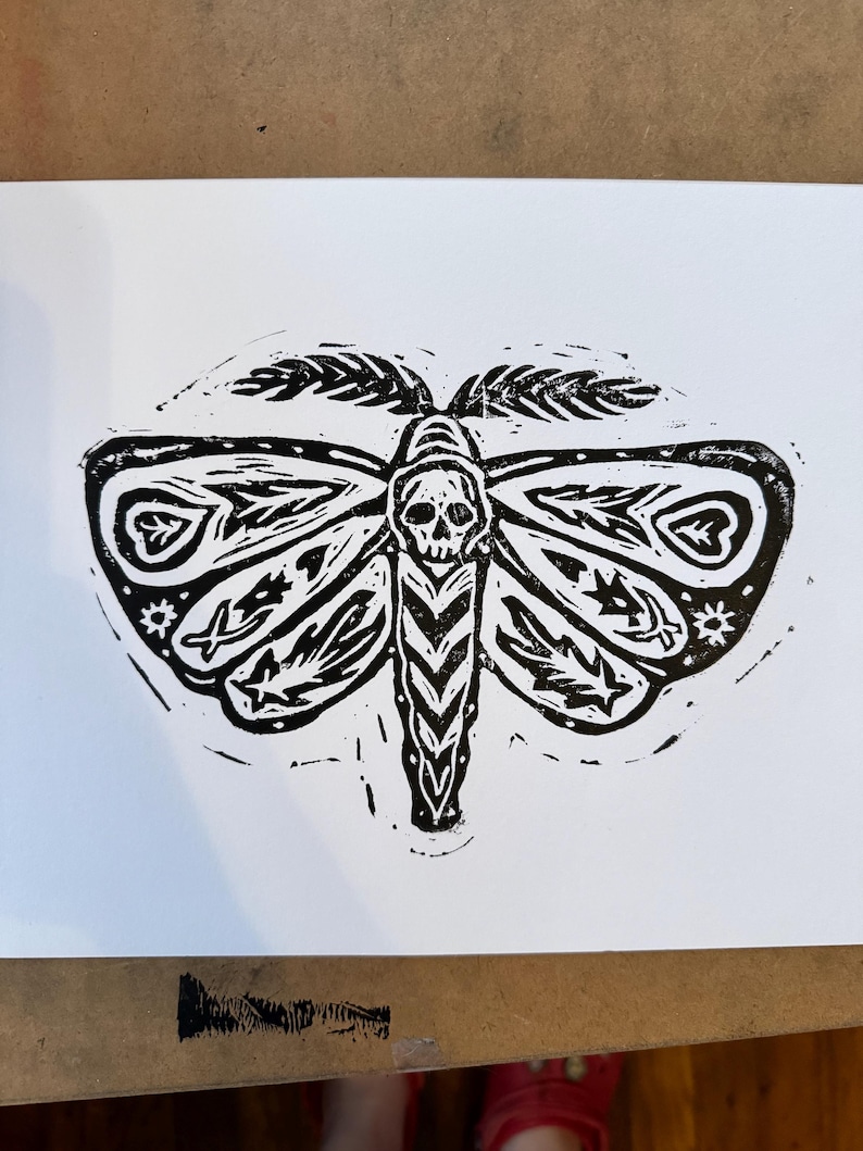 May include: Black and white linocut print of a moth with a skull in its body. The moth has intricate details and patterns on its wings.