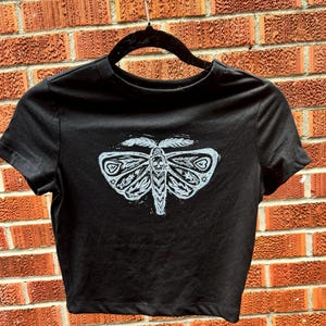 May include: Black cropped t-shirt with a white graphic of a moth with intricate details.