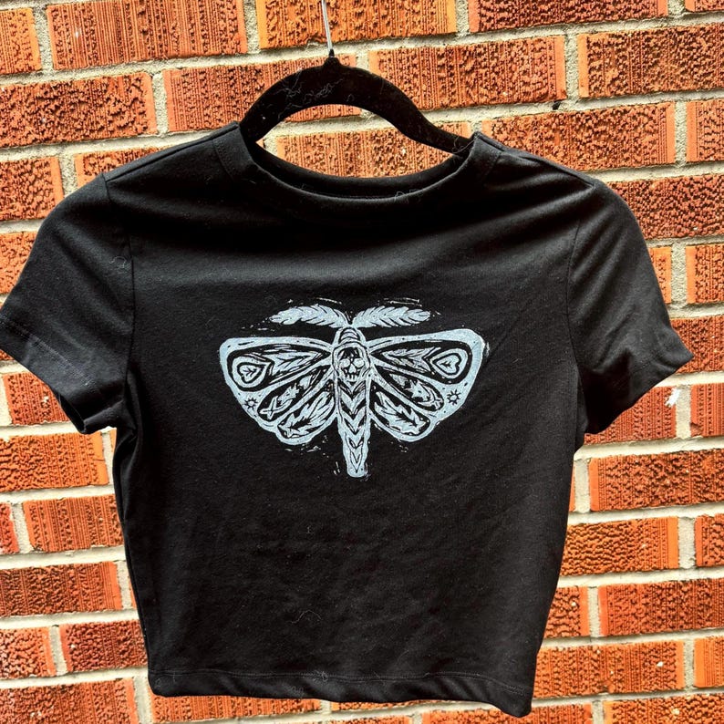 May include: Black cropped t-shirt with a white graphic of a moth with intricate details.