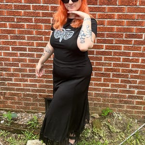 May include: A woman wearing a black t-shirt with a white moth graphic and a black maxi skirt with lace trim. She is wearing black sunglasses and has bright orange hair.