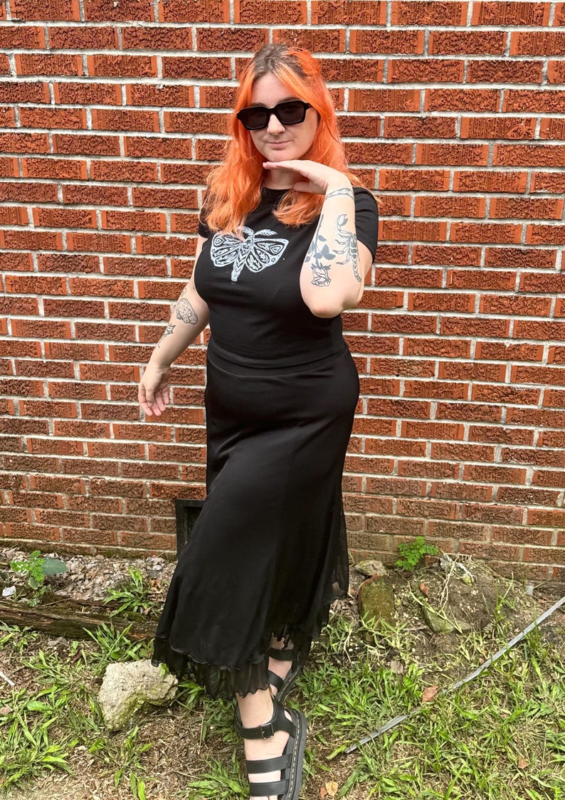 May include: A woman wearing a black t-shirt with a white moth graphic and a black maxi skirt with lace trim. She is wearing black sunglasses and has bright orange hair.