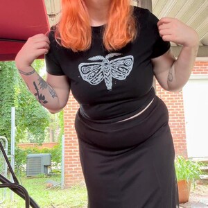 May include: A black cropped t-shirt with a white moth graphic. The shirt is paired with a black skirt.