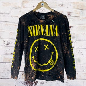 Puede incluir: Camiseta negra de manga larga con detalles desgastados y un gráfico de carita sonriente amarilla de Nirvana. La palabra "NIRVANA" está impresa en letras amarillas en bloque sobre la carita sonriente. Las mangas tienen texto amarillo.