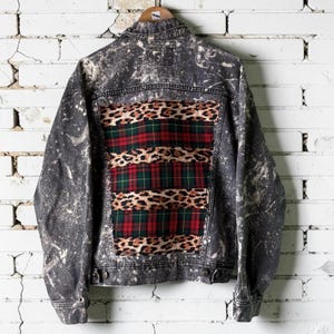 May include: A charcoal gray denim jacket featuring a bleached splatter pattern throughout. The back panel displays a rectangular insert composed of alternating horizontal stripes of leopard print fabric and red and green plaid fabric.