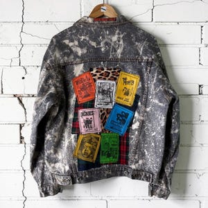 May include: A grey acid-wash denim jacket featuring a back panel decorated with a collage of colorful rectangular patches. The patches include band names like The Dickies, The Misfits, and Ramones, alongside leopard print and plaid fabric accents.