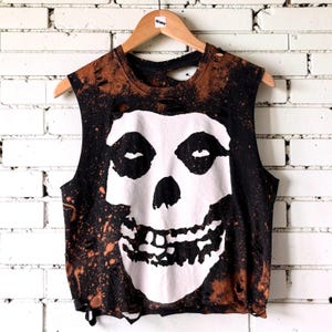 May include: Black and brown sleeveless crop top with a white skull graphic. The shirt has a distressed, bleached design with holes and a raw hem. The skull graphic is centered on the front.