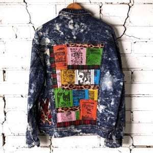 May include: A blue acid-wash denim jacket featuring a back panel decorated with a collage of colorful band patches, including Ramones, Black Flag, and The Cramps, separated by strips of leopard print fabric and red and green plaid fabric.