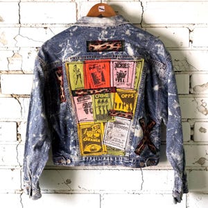 May include: A blue acid-wash denim jacket featuring a collage of colorful rectangular band posters on the back. The patches include text such as Poison Idea, The Dickies, Bad Brains, Crime, Offs, Agent Orange, and Descendents, accented by leopard print strips.