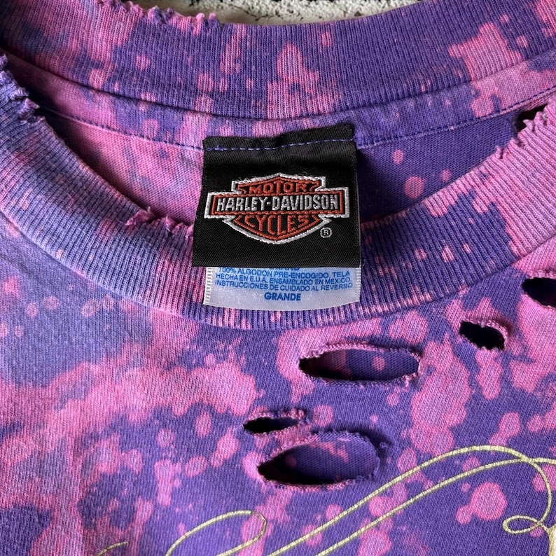 May include: Close-up of a purple and pink Harley-Davidson Cycles t-shirt. The shirt has a distressed look with holes and a faded tie-dye pattern. The Harley-Davidson label is visible.