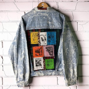 May include: A light blue acid-wash denim jacket featuring a back panel with a dark green plaid fabric overlay. The panel displays six rectangular patches with band names and graphics including Black Flag, Poison Idea, Bad Religion, and The Cramps.