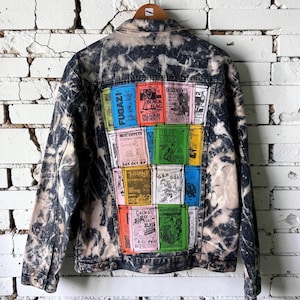May include: A black and white acid-wash denim jacket featuring a collage of colorful rectangular punk rock concert posters on the back. The patches include text such as Fugazi, Black Flag, Descendents, Minor Threat, G.B.H, and Circle Jerks.