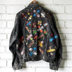 May include: A black denim jacket featuring a distressed, acid-wash finish with white paint splatters. The back is decorated with numerous rectangular patches in various colors and patterns, including plaid, leopard print, and solid hues.