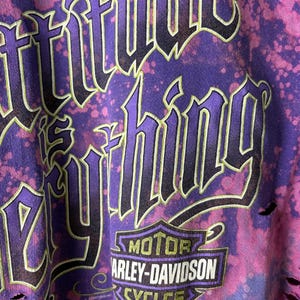 May include: Purple and pink distressed t-shirt with the words "Attitude is Everything" in a gothic font. Features the Harley-Davidson logo at the bottom. The shirt has a tie-dye effect and distressed edges.