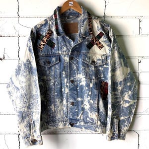 May include: A blue denim jacket featuring a white bleach-splatter pattern, a brown leather collar tag, and decorative cross-shaped patches on the chest. The patches combine leopard print fabric with dark green and red plaid tartan patterns.
