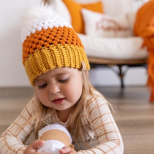 May include: A young child wearing a crocheted orange, white, and yellow hat with a white pom pom on top. The child is holding a white and brown object in their hands.
