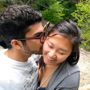 May include: A man and a woman are standing close together in a natural setting. The man is kissing the woman on the cheek. The woman is smiling and looking down. The man is wearing a white t-shirt with a star pattern and the woman is wearing a grey hooded sweatshirt.