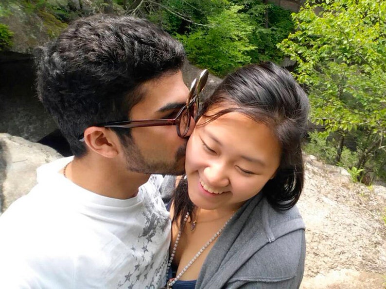 May include: A man and a woman are standing close together in a natural setting. The man is kissing the woman on the cheek. The woman is smiling and looking down. The man is wearing a white t-shirt with a star pattern and the woman is wearing a grey hooded sweatshirt.