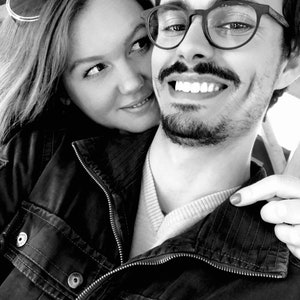 May include: A black and white photo of a couple smiling. The man is wearing a black jacket and glasses. The woman is wearing a jacket and sunglasses.