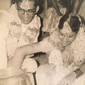 May include: A black and white photo of a wedding ceremony. The bride and groom are wearing traditional Indian clothing and garlands of flowers. The bride is smiling and looking down at her hands.