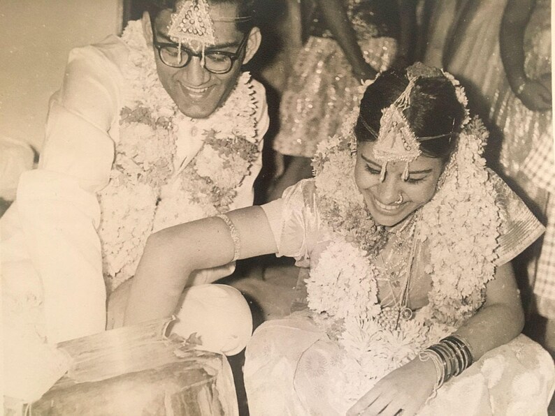 May include: A black and white photo of a wedding ceremony. The bride and groom are wearing traditional Indian clothing and garlands of flowers. The bride is smiling and looking down at her hands.