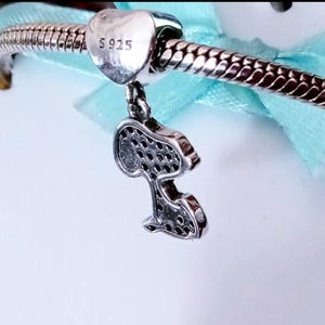 May include: A silver charm bracelet featuring a heart-shaped bead stamped with S925 and a dangling pendant shaped like a cartoon dog silhouette. The pendant is covered in small, dark, circular indentations across its surface, reflecting light.