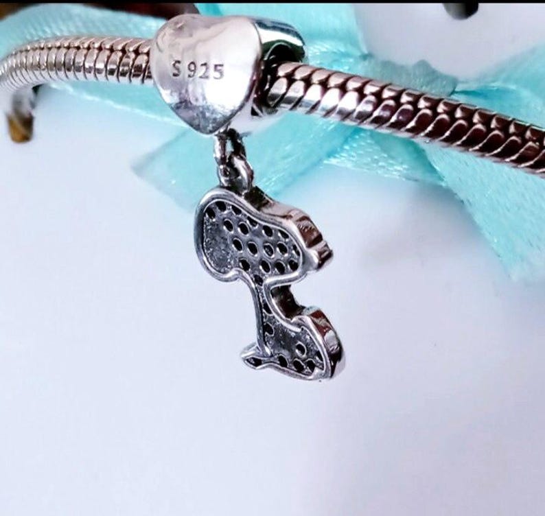 May include: A silver charm bracelet featuring a heart-shaped bead stamped with S925 and a dangling pendant shaped like a cartoon dog silhouette. The pendant is covered in small, dark, circular indentations across its surface, reflecting light.