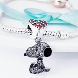 May include: A silver charm bracelet featuring a dangling Snoopy character pendant encrusted with clear cubic zirconia stones and a black enamel nose. Above the pendant is a heart-shaped charm set with numerous small red stones.