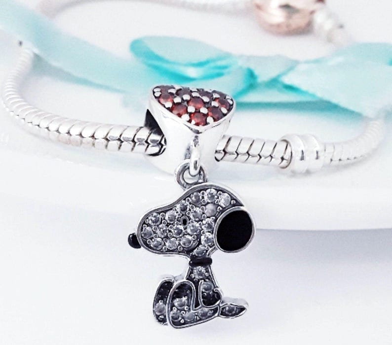 May include: A silver charm bracelet featuring a dangling Snoopy character pendant encrusted with clear cubic zirconia stones and a black enamel nose. Above the pendant is a heart-shaped charm set with numerous small red stones.