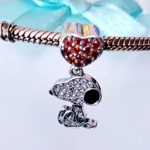 May include: A silver-toned charm bracelet bead featuring a heart-shaped top encrusted with small red stones. Suspended from the heart is a silver-toned charm shaped like a cartoon dog, covered in clear white crystals with a black enamel nose.
