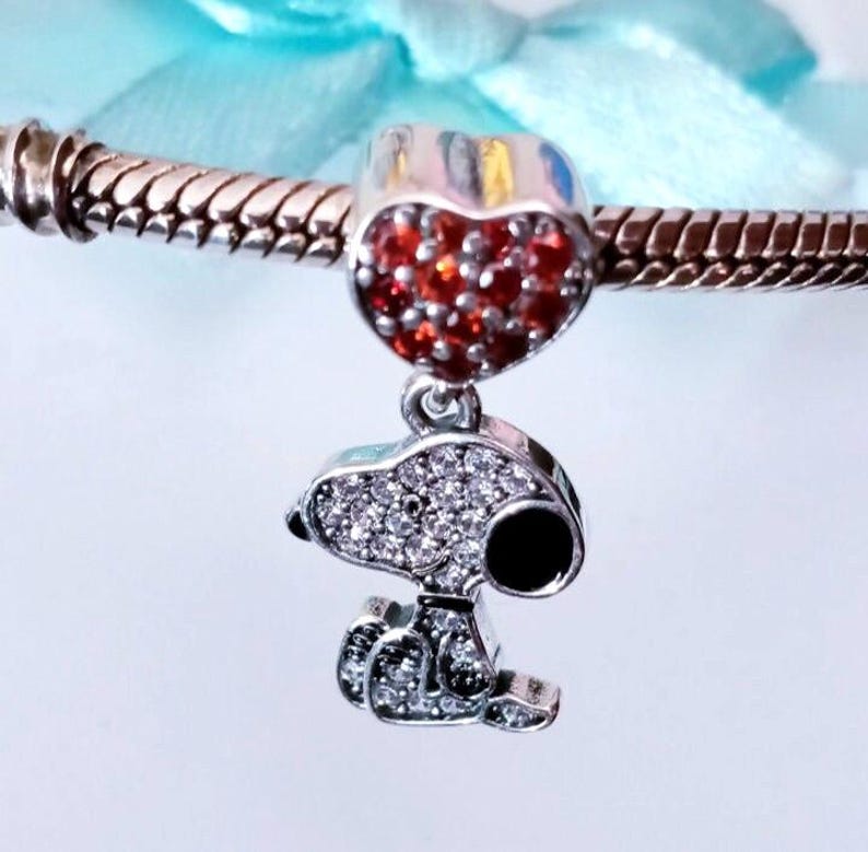 May include: A silver-toned charm bracelet bead featuring a heart-shaped top encrusted with small red stones. Suspended from the heart is a silver-toned charm shaped like a cartoon dog, covered in clear white crystals with a black enamel nose.