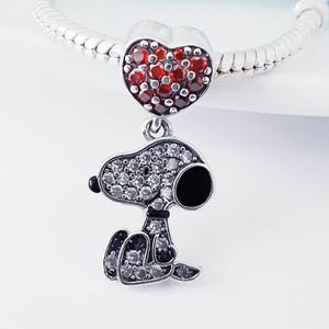 May include: A silver charm bracelet bead featuring a heart-shaped charm encrusted with red stones, attached to a dangling pendant shaped like a cartoon dog. The dog pendant is covered in clear white stones with a black enamel ear and nose detail.