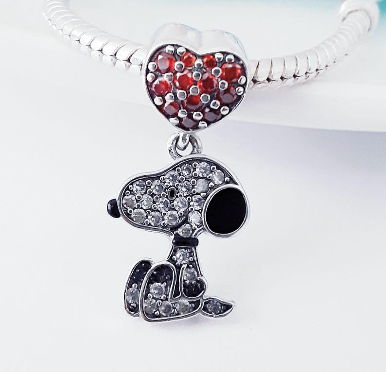 May include: A silver charm bracelet bead featuring a heart-shaped charm encrusted with red stones, attached to a dangling pendant shaped like a cartoon dog. The dog pendant is covered in clear white stones with a black enamel ear and nose detail.