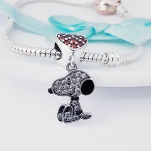 May include: A silver charm bracelet featuring a dangling Snoopy pendant encrusted with clear cubic zirconia stones and a black enamel ear. The bracelet includes a heart-shaped charm paved with red stones and a rose gold-toned clasp component.
