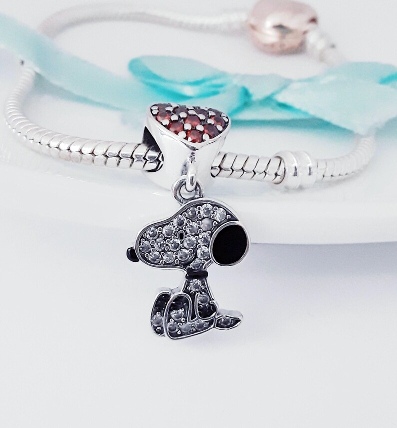 May include: A silver charm bracelet featuring a dangling Snoopy pendant encrusted with clear cubic zirconia stones and a black enamel ear. The bracelet includes a heart-shaped charm paved with red stones and a rose gold-toned clasp component.