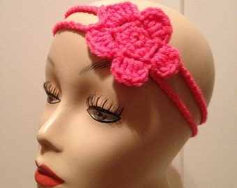May include: A pink crocheted headband with a flower design. The headband is worn on a mannequin head.