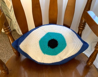 May include: A crocheted pillow in the shape of a blue evil eye. The eye is white with a blue iris and a black pupil.