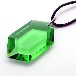 Pode incluir: Um pingente de cristal verde esmeralda facetado com um cordão preto. O pingente tem uma forma geométrica com múltiplas facetas que captam a luz. É suspenso por um laço de metal prateado preso a um cordão preto.
