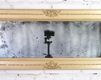 May include: A rectangular wall mirror featuring an ornate, metallic gold frame with detailed corner carvings. The mirror glass has a distressed, mottled finish with dark spots and a cloudy appearance, reflecting a camera mounted on a tripod.