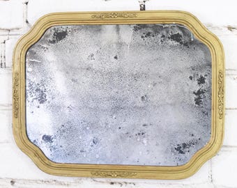 May include: A rectangular mirror with a gold-toned frame featuring decorative embossed floral details at the center of each side. The glass surface displays significant age-related oxidation, characterized by dark, mottled spots and a weathered finish.