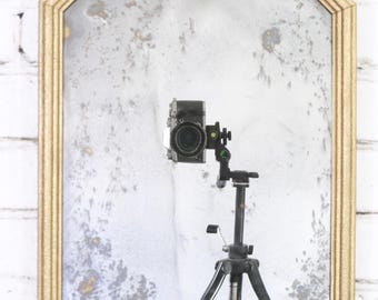 May include: A rectangular wall mirror with a gold-toned frame featuring clipped top corners. The glass surface displays a distressed, mottled finish with grey and white oxidation patterns, reflecting a camera mounted on a black tripod.