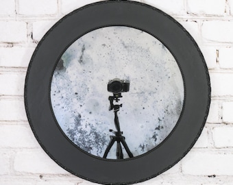 May include: A round wall mirror featuring a wide, matte black frame with a subtle beaded edge detail. The glass surface has a distressed, mottled finish with dark gray and white spots, reflecting a camera mounted on a tripod in the center.