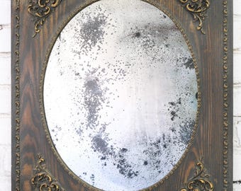 May include: A rectangular wall mirror with a dark stained wood frame featuring an oval center opening. The frame is decorated with gold-toned ornate corner accents and a beaded inner border. The glass surface shows significant silvering oxidation.