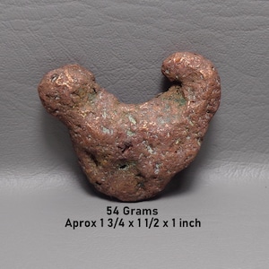 May include: A raw copper nugget with a textured, reddish-brown surface and green oxidation spots. The object weighs 54 grams and measures approximately 1 3/4 by 1 1/2 by 1 inches, as indicated by the printed text below the specimen.