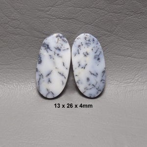 May include: Two oval-shaped white gemstones with black dendritic inclusions. The stones measure 13 x 26 x 4mm. The gemstones have a polished surface and are set against a gray background.