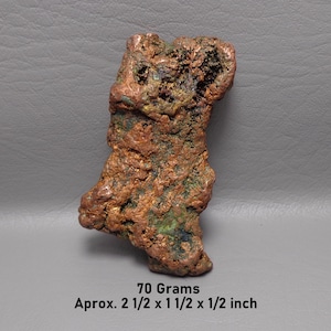 May include: A raw copper nugget with a textured, metallic surface featuring reddish-brown and green oxidation. The specimen weighs 70 grams and measures approximately 2 1/2 x 1 1/2 x 1/2 inches as indicated by the text printed below the object.