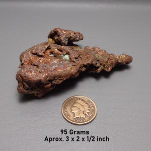 May include: A raw copper nugget with a textured, metallic brown surface and hints of green oxidation. Next to it is a 1903 United States Indian Head penny for scale. The specimen weighs 95 grams and measures approximately 7.6 x 5.1 x 1.3 cm.